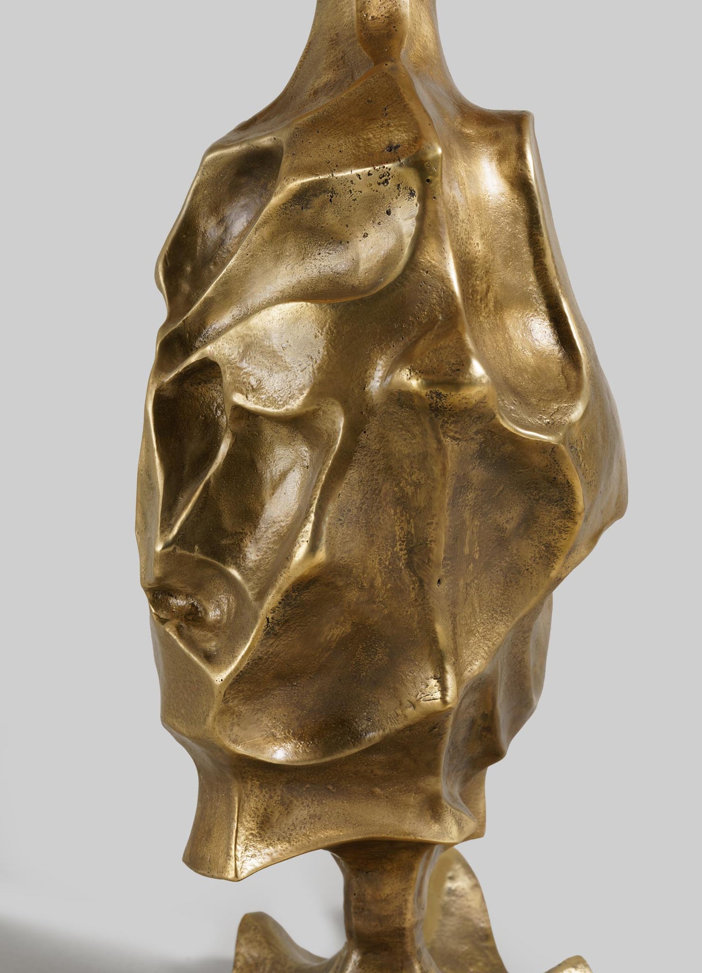 The VIYA Indian Vessel Table Lamp features an abstract bronze design with flowing curves and a textured metallic surface, inspired by traditional surahi vessels. Its organic form casts dynamic shadows against a plain gray background.