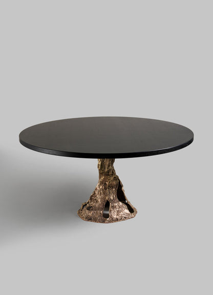 The VIYA Kikar Dining Table features a round dark wood top and a sculpted bronze base inspired by a Kikar tree trunk, from the Desert Collection, shown against a plain light gray background.