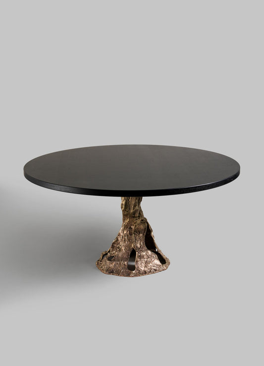 The VIYA Kikar Dining Table features a round dark wood top and a sculpted bronze base inspired by a Kikar tree trunk, from the Desert Collection, shown against a plain light gray background.