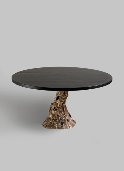 The VIYA Kikar Dining Table features a round black top and a sculptural metallic base inspired by the Kikar tree’s abstract, textured roots, shown against a plain gray background.