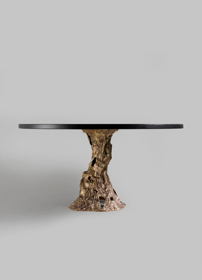 The VIYA Kikar Dining Table features a smooth black round top and a sculptural gold base inspired by the Kikar tree, showcasing organic elegance against a plain light gray background.