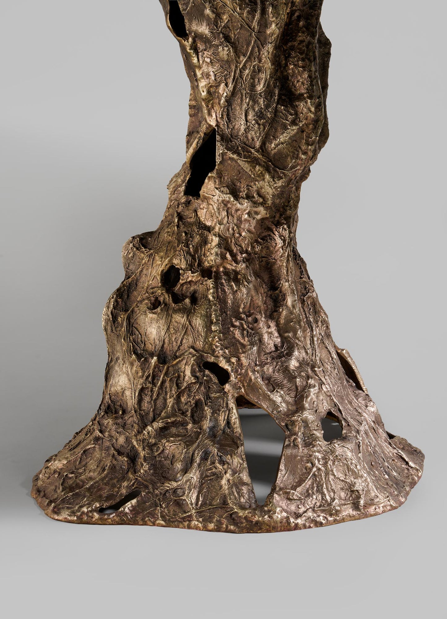 The VIYA Kikar Dining Table features a bronze base with a textured, organic surface and irregular contours inspired by the Kikar tree. Hollowed openings evoke roots or trunks, creating a desert-inspired design set against a gray background.