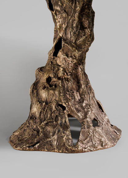 The VIYA Kikar Dining Table features a bronze base with a textured, organic surface and irregular contours inspired by the Kikar tree. Hollowed openings evoke roots or trunks, creating a desert-inspired design set against a gray background.