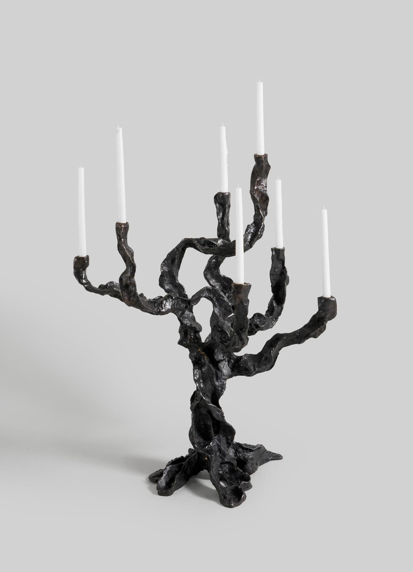 The VIYA Meghalaya Candelabra Large is a sculptural black metal piece inspired by living root bridges, with twisted branch-like arms holding seven tall white candles, set against a plain grey background.