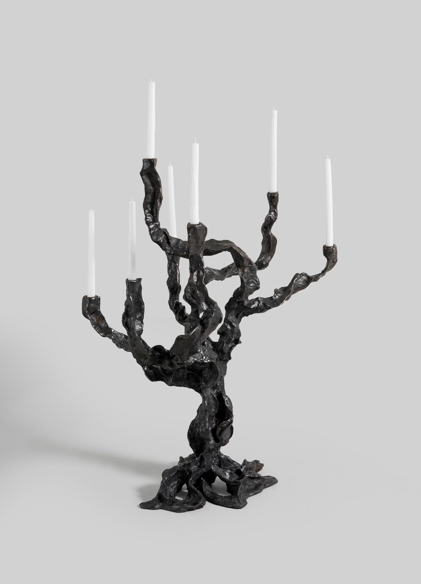 The Meghalaya Candelabra Large by VIYA features a sculptural dark metal design inspired by living root bridges, with a twisted, organic shape holding seven white taper candles.
