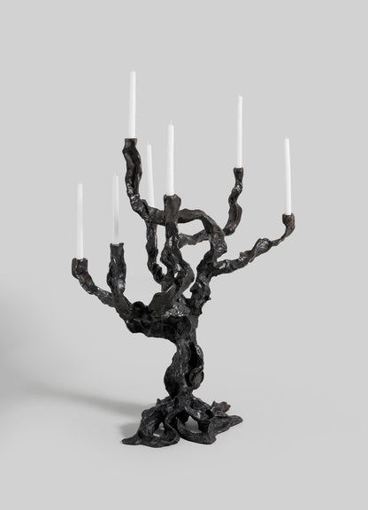 The Meghalaya Candelabra Large by VIYA features a sculptural dark metal design inspired by living root bridges, with a twisted, organic shape holding seven white taper candles.