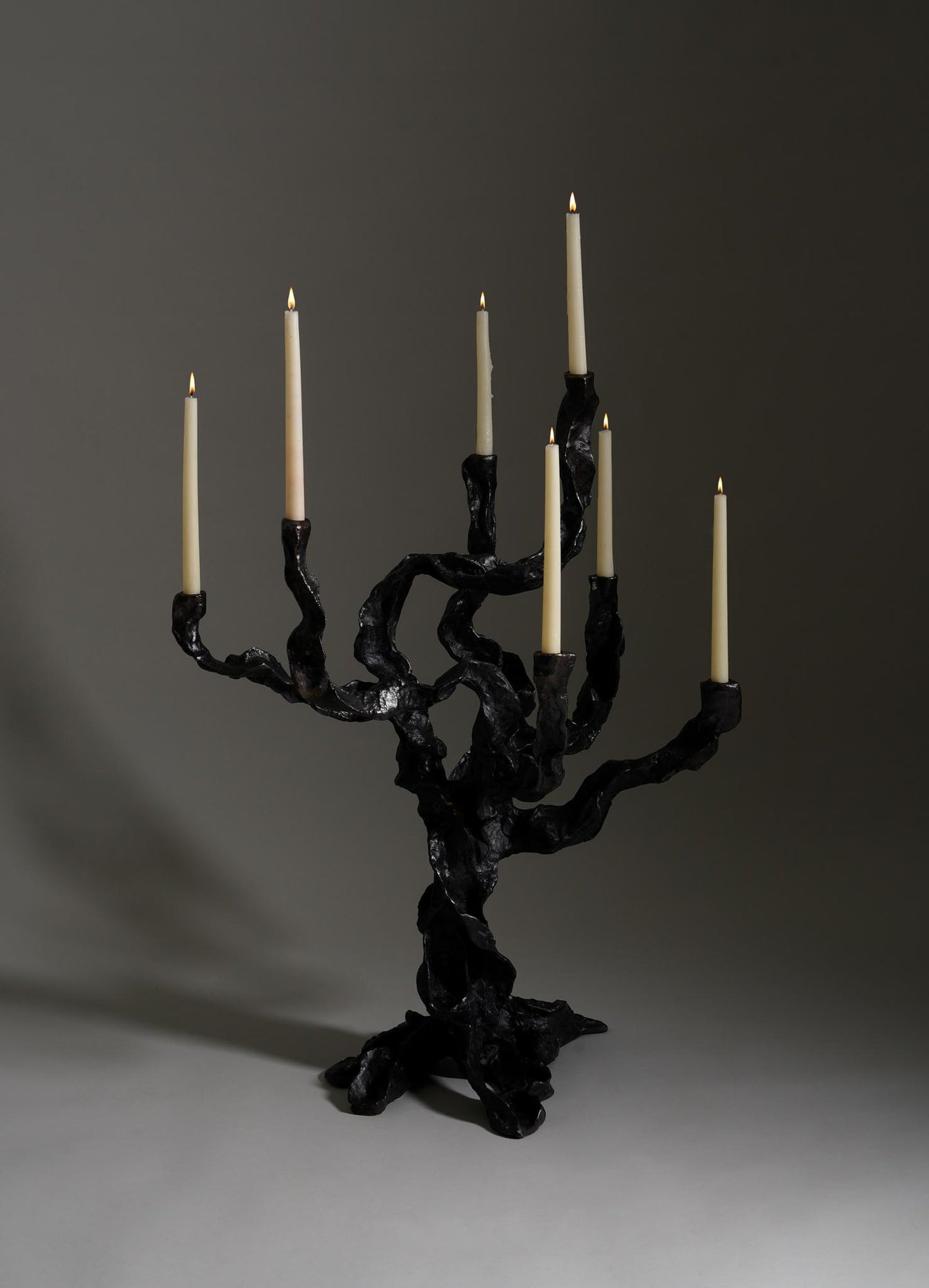 The Meghalaya Candelabra Large by VIYA, inspired by living root bridges, displays seven lit tapers. Its black, sculptural form adds an organic accent to brass decor against a plain gray background.
