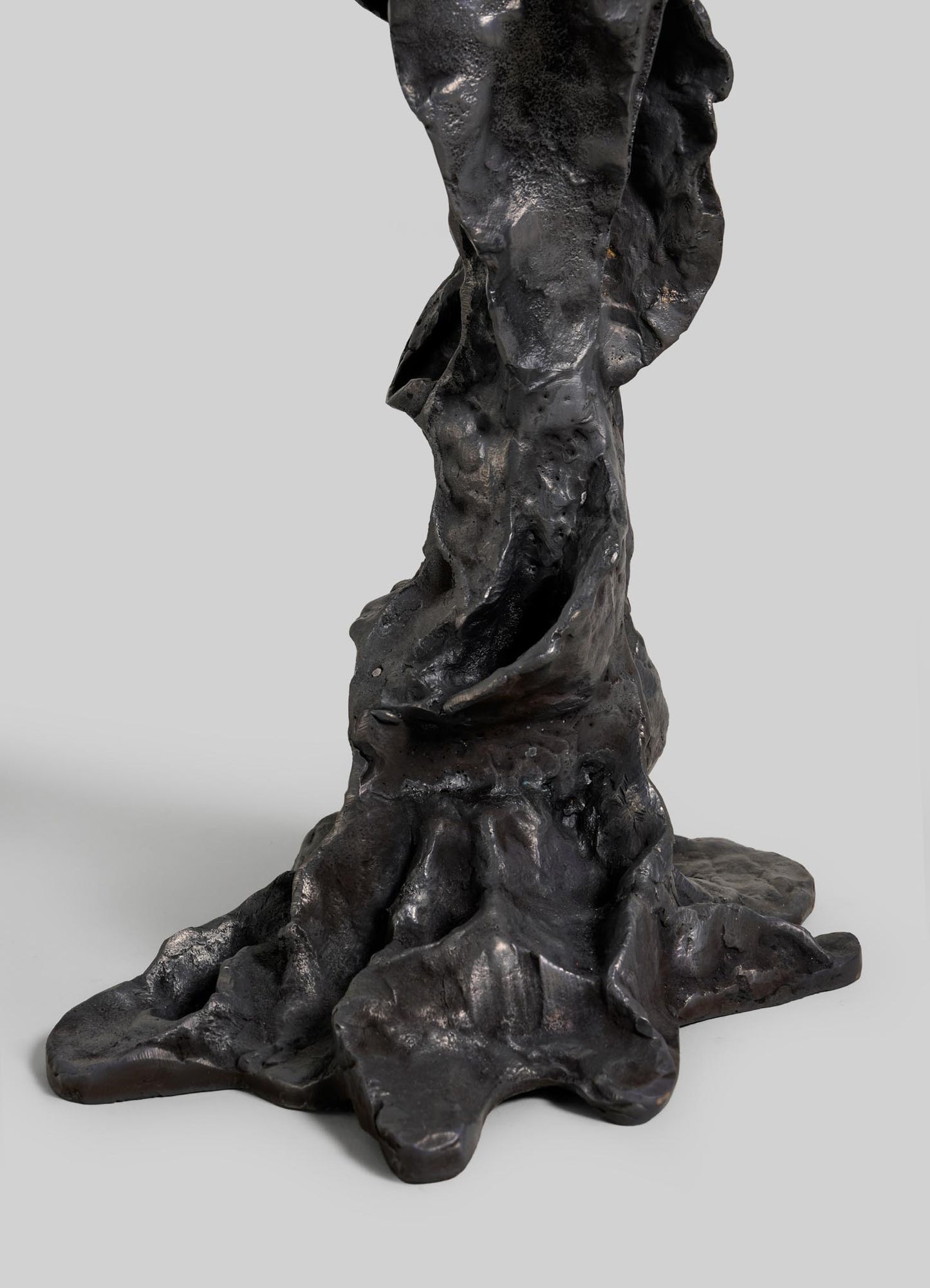 A close-up of the Meghalaya Candelabra Large by VIYA shows its dark, textured bronze base with organic, uneven shapes reminiscent of living root bridges, resembling tree roots or molten material against a plain gray background.