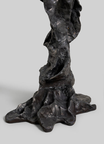 A close-up of the Meghalaya Candelabra Large by VIYA shows its dark, textured bronze base with organic, uneven shapes reminiscent of living root bridges, resembling tree roots or molten material against a plain gray background.