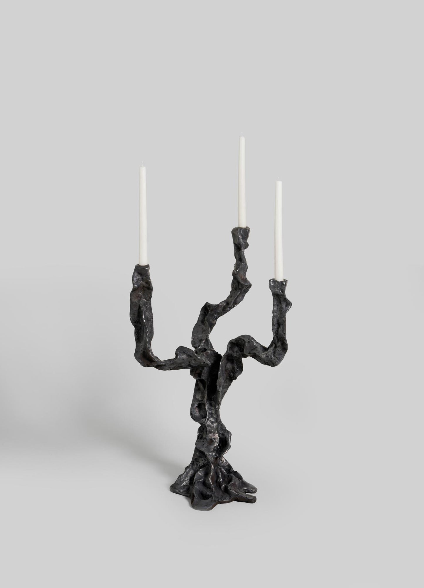 The VIYA Meghalaya Candelabra Small is a sculptural black metal piece with three twisting, root-bridge-inspired arms, each holding a tall white taper candle, set against a plain light gray background.