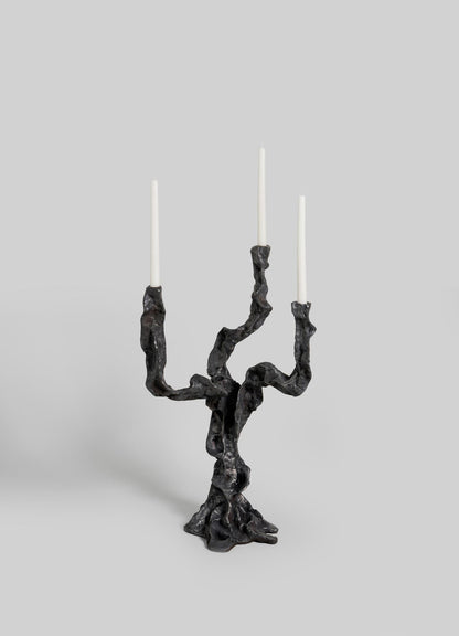The VIYA Meghalaya Candelabra Small is a sculptural black metal piece with three twisting, root-bridge-inspired arms, each holding a tall white taper candle, set against a plain light gray background.