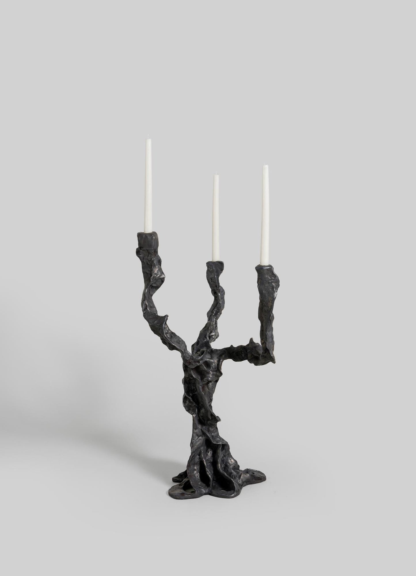 The VIYA Meghalaya Candelabra Small features three twisted, root bridge-inspired black metal branches holding slender white candles, displayed against a plain light gray background.