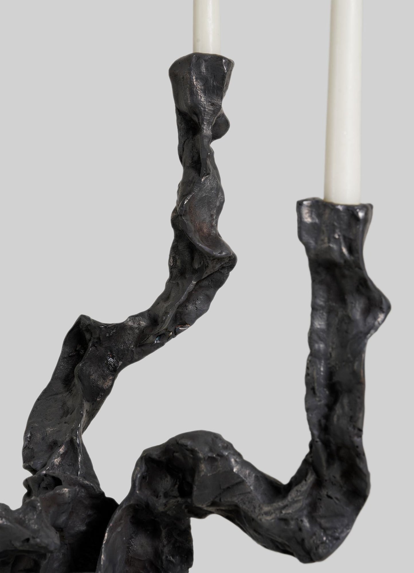 A close-up of VIYA's Meghalaya Candelabra Small, a black metal piece with a twisted, textured design inspired by living root bridges, holds two white taper candles against a plain gray background.