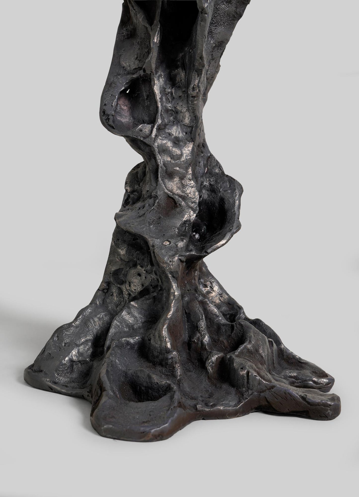 A close-up of the Meghalaya Candelabra Small by VIYA shows its abstract, dark metallic form with an organic, textured surface—like twisted rock or root bridges—set against a plain light gray background.