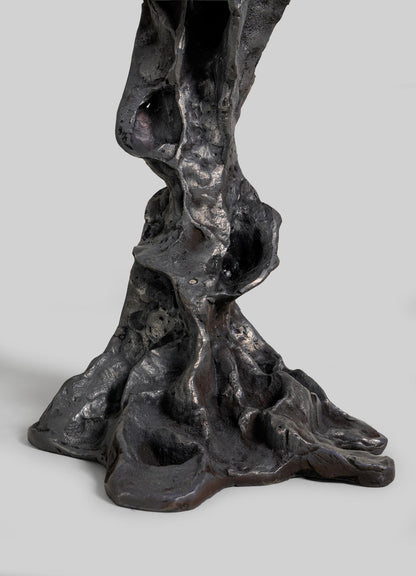 A close-up of the Meghalaya Candelabra Small by VIYA shows its abstract, dark metallic form with an organic, textured surface—like twisted rock or root bridges—set against a plain light gray background.