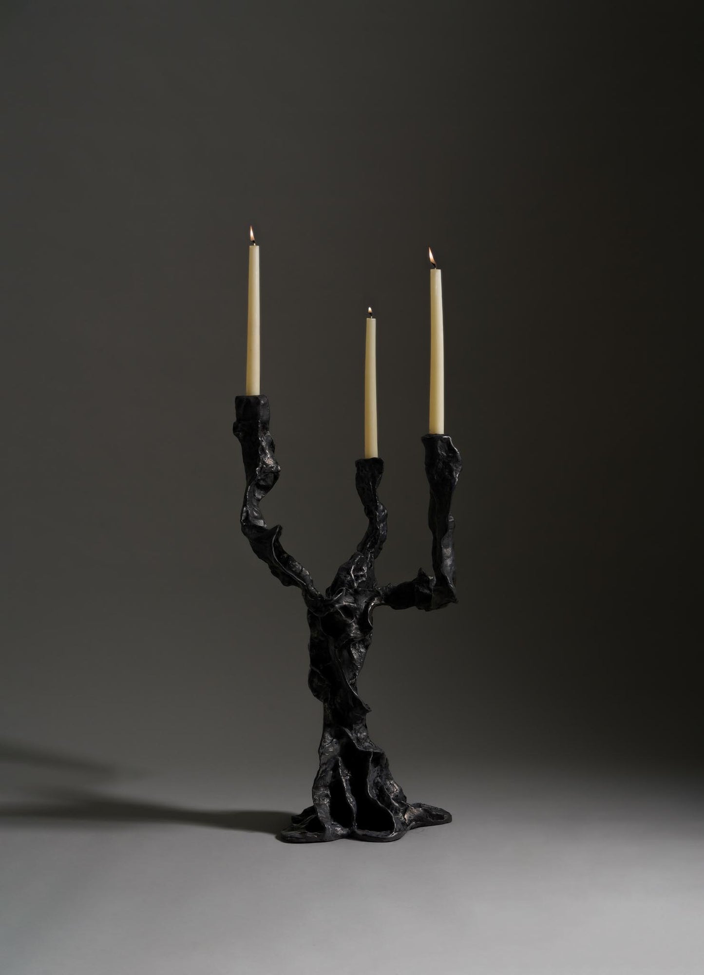 The VIYA Meghalaya Candelabra Small features three twisted black arms inspired by living root bridges, each holding a tall beige candle. Its sculptural, molten look stands out against a dark background.
