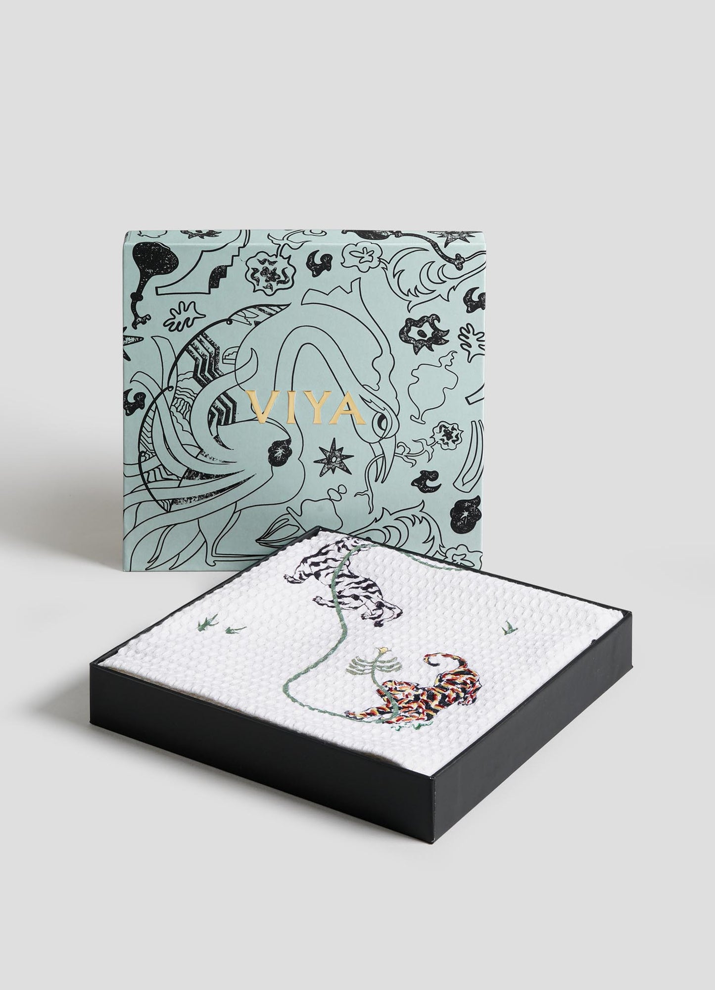 The Lounging Tiger Hand Towels - Set of 2 by Viya are displayed in a decorative box with floral and tiger designs, alongside a matching tray featuring two tigers and a flower, set on a white textured surface against a light background.