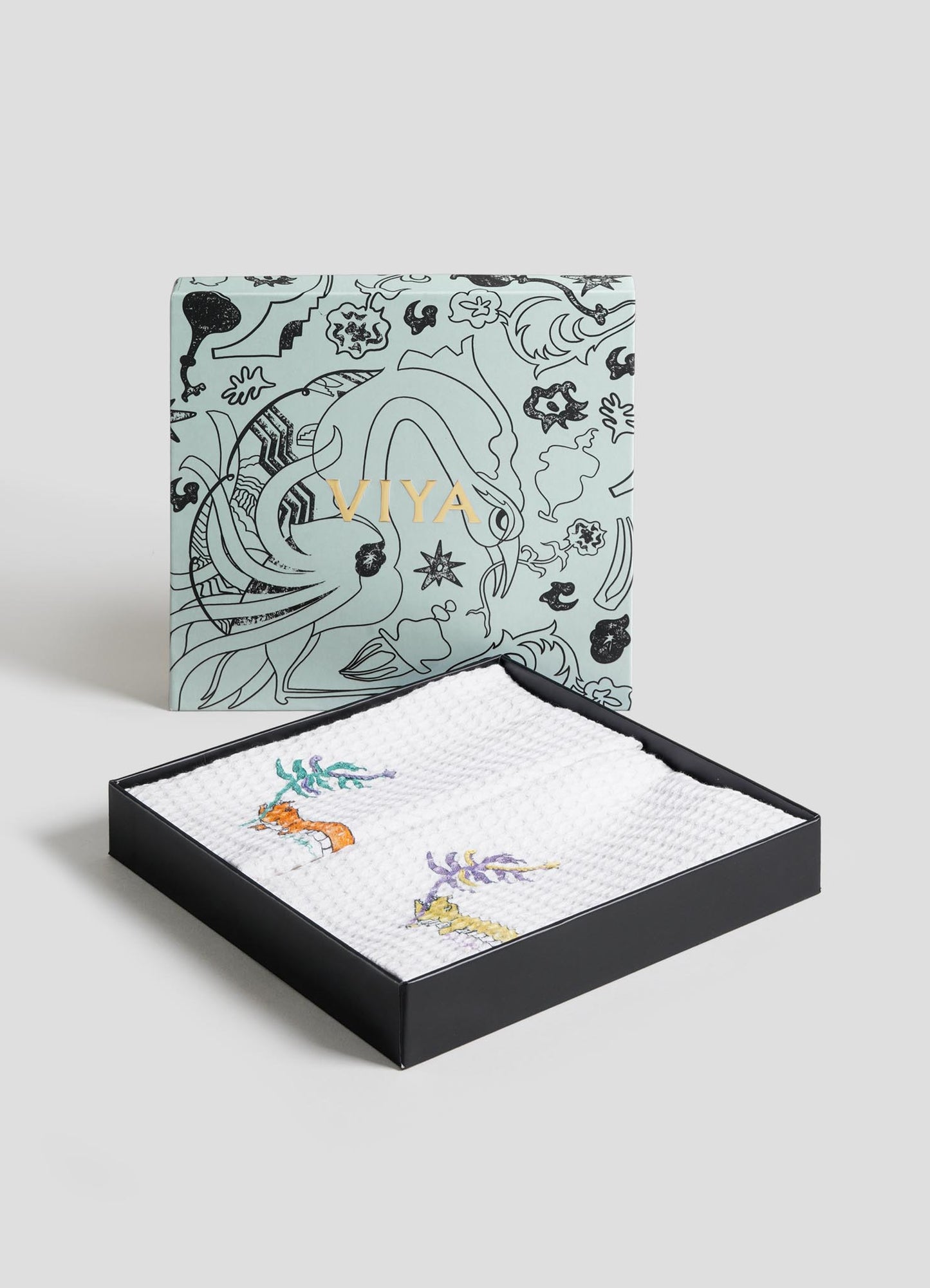 The Lurking Crocodile Hand Towels by Viya come as a set of 2, packaged in a decorative black box with "Viya" in gold, featuring folded white towels embroidered with vibrant aquatic animals and plants.