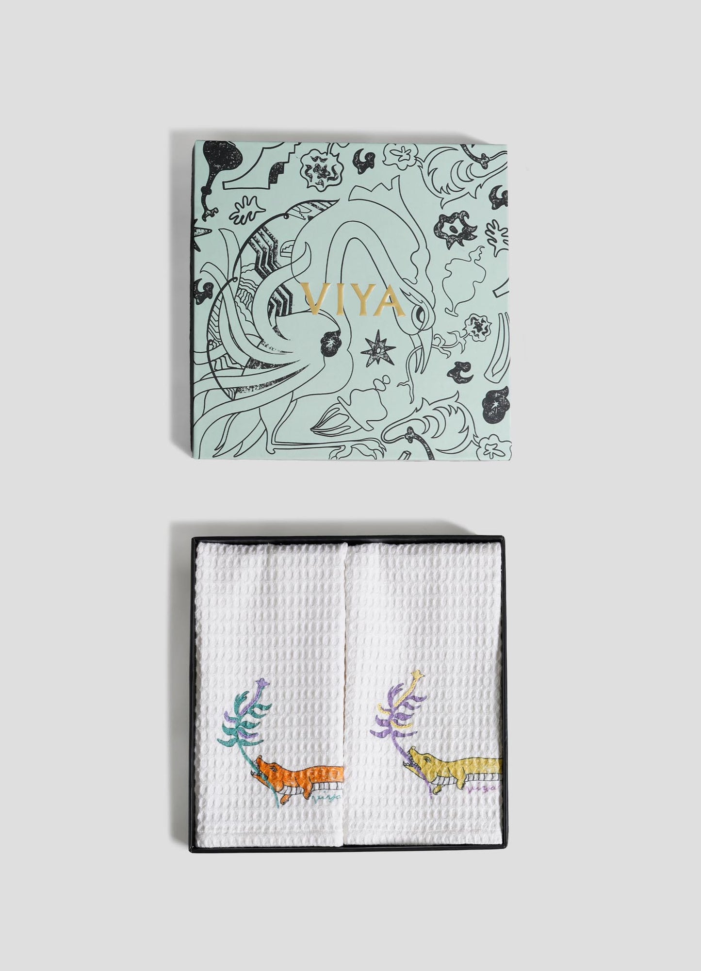 A light blue Viya box with black animal and plant designs and gold text sits atop white textured Lurking Crocodile Hand Towels—set of 2—embroidered with vibrant animals, capturing Indian wildlife in a jungle-inspired home decor style.