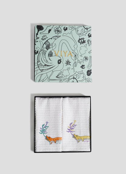 A light blue Viya box with black animal and plant designs and gold text sits atop white textured Lurking Crocodile Hand Towels—set of 2—embroidered with vibrant animals, capturing Indian wildlife in a jungle-inspired home decor style.