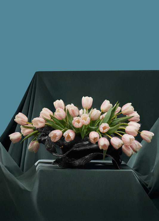 A floral arrangement of pale pink tulips in the VIYA Leher Vessel sits on a table draped with dark green Leheriya textile, set against a solid teal background.