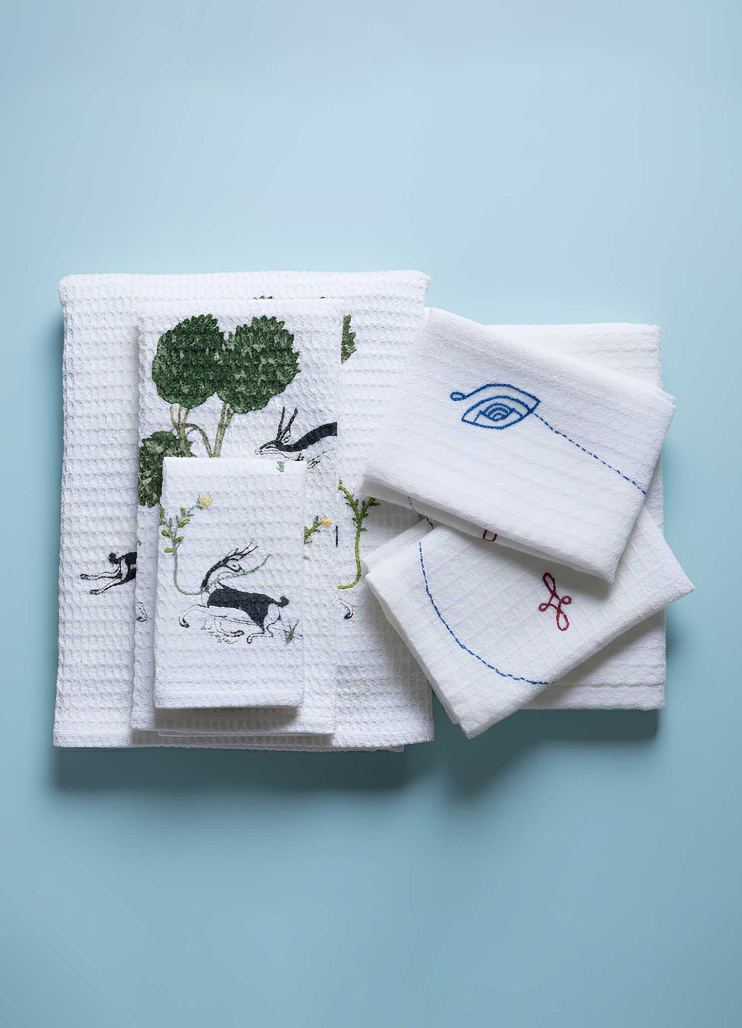 A set of Viya Majestic Blackbuck Face Towels (4 pcs) featuring white textured fabric with colorful embroidery inspired by Indian wildlife art—showcasing animals, plants, and abstract motifs—displayed on a light blue surface.