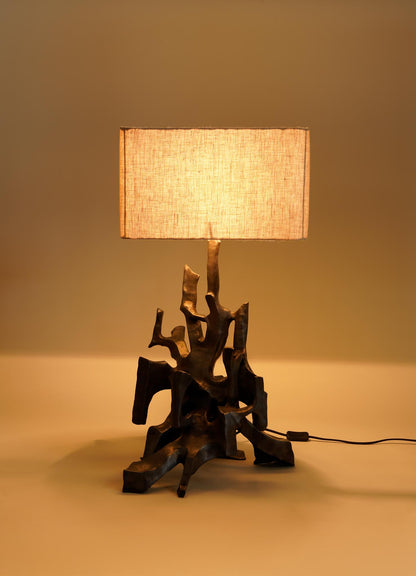 The VIYA Maski Table Lamp features a sculptural abstract bronze base and a rectangular beige fabric shade, casting a warm glow as it sits on a plain surface.