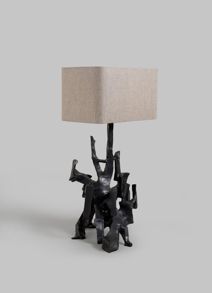 The Maski Table Lamp by VIYA features an abstract black metal base resembling intertwined figures and a rectangular textured beige lampshade, displayed against a plain gray background.