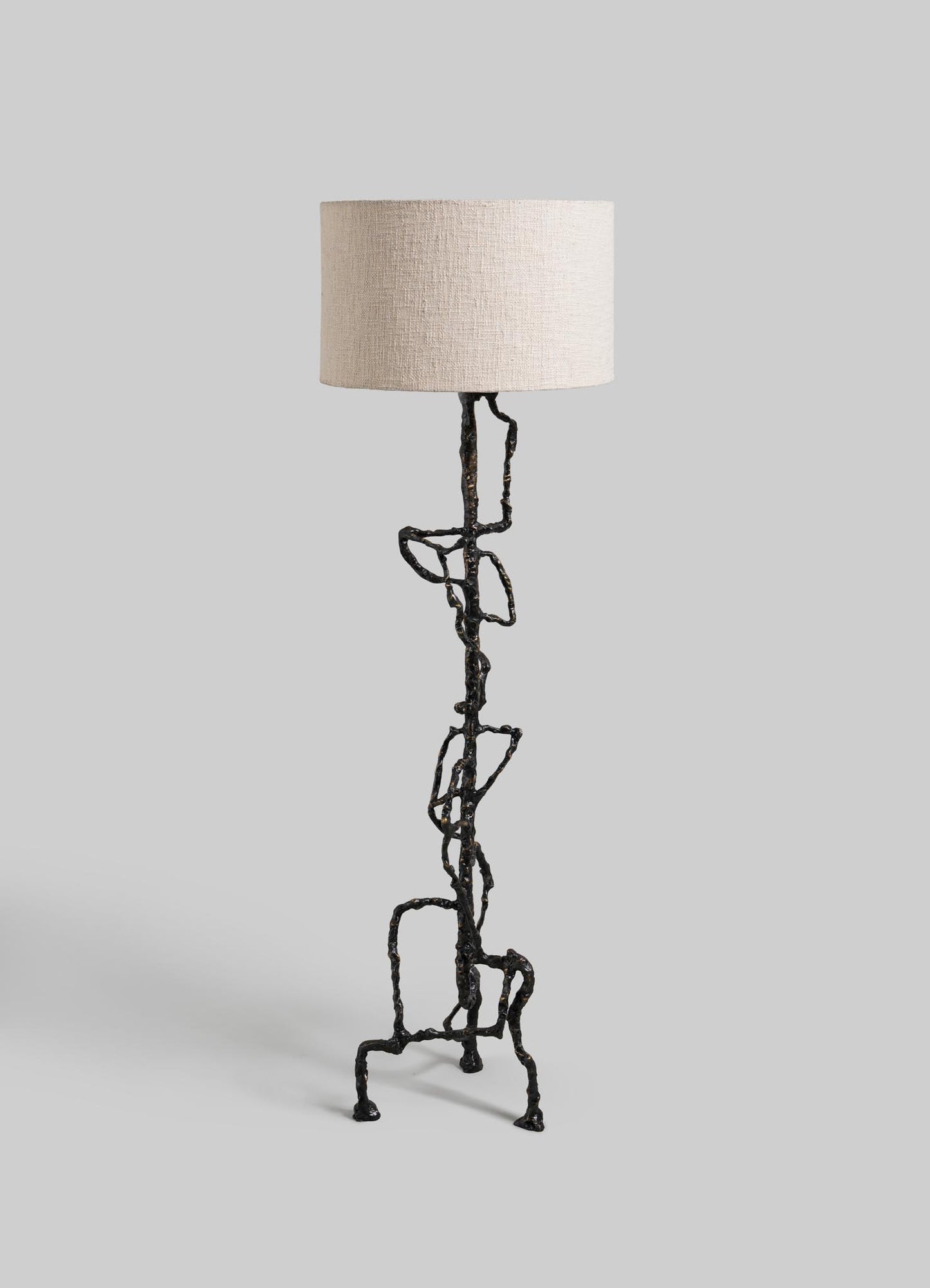 The VIYA Mohenjo Daro Floor Lamp features a beige fabric shade and an artistic, twisted black metal base inspired by abstract forms, evoking the lost civilization of Mohenjo-Daro, set against a plain light gray background.