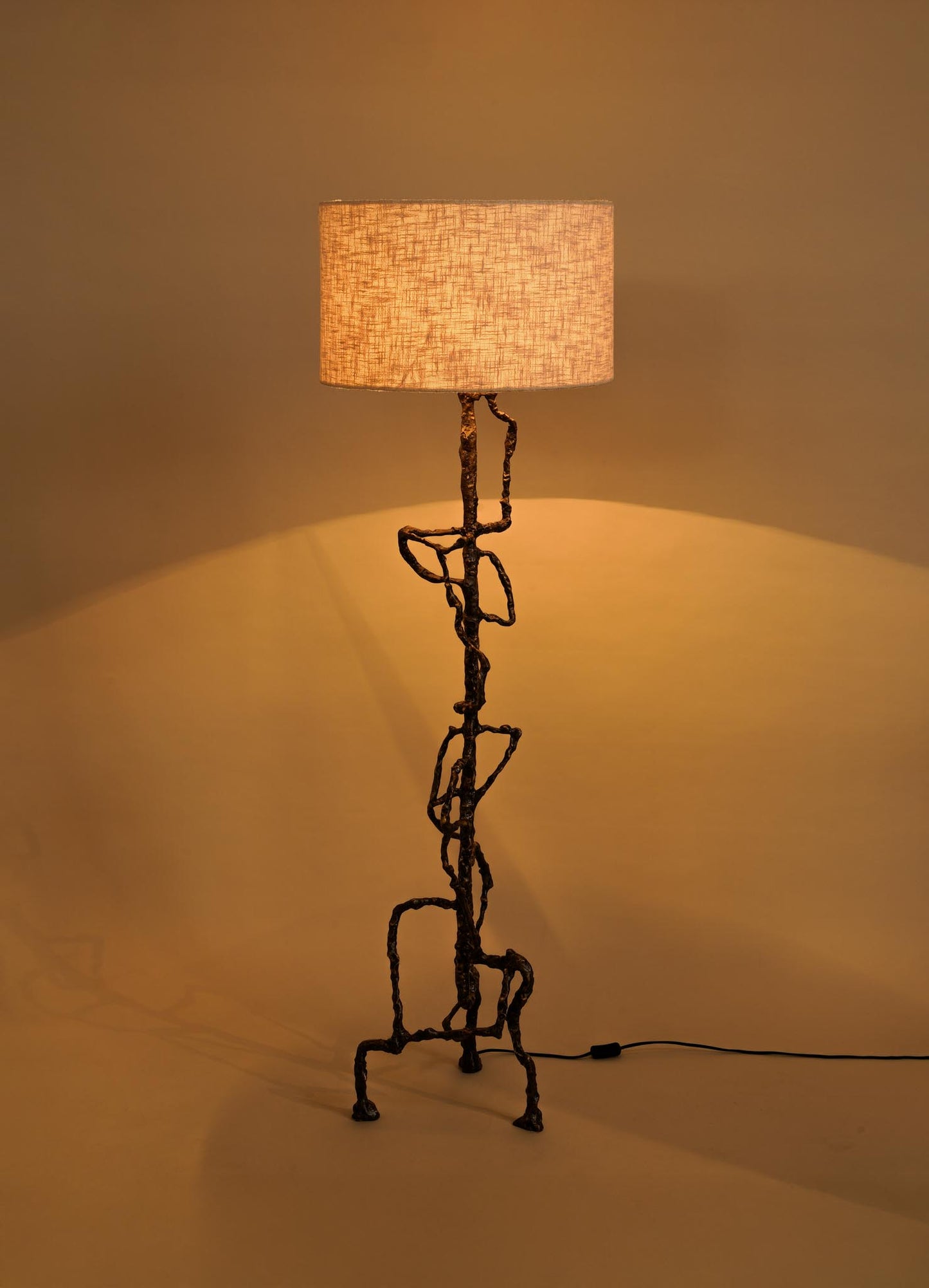 The VIYA Mohenjo Daro Floor Lamp features a textured, sculptural metal base inspired by Indus Valley art and a cylindrical beige fabric shade, casting a warm glow against a plain beige background.