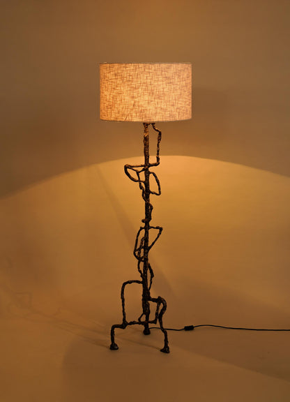 The VIYA Mohenjo Daro Floor Lamp features a textured, sculptural metal base inspired by Indus Valley art and a cylindrical beige fabric shade, casting a warm glow against a plain beige background.