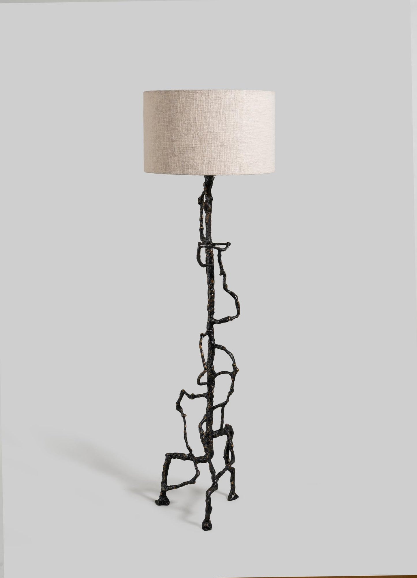 The Mohenjo Daro Floor Lamp by VIYA features an abstract, branching bronze-finished metal base inspired by Indus Valley art and a simple beige fabric shade, set against a plain gray background.