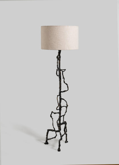 The Mohenjo Daro Floor Lamp by VIYA features an abstract, branching bronze-finished metal base inspired by Indus Valley art and a simple beige fabric shade, set against a plain gray background.
