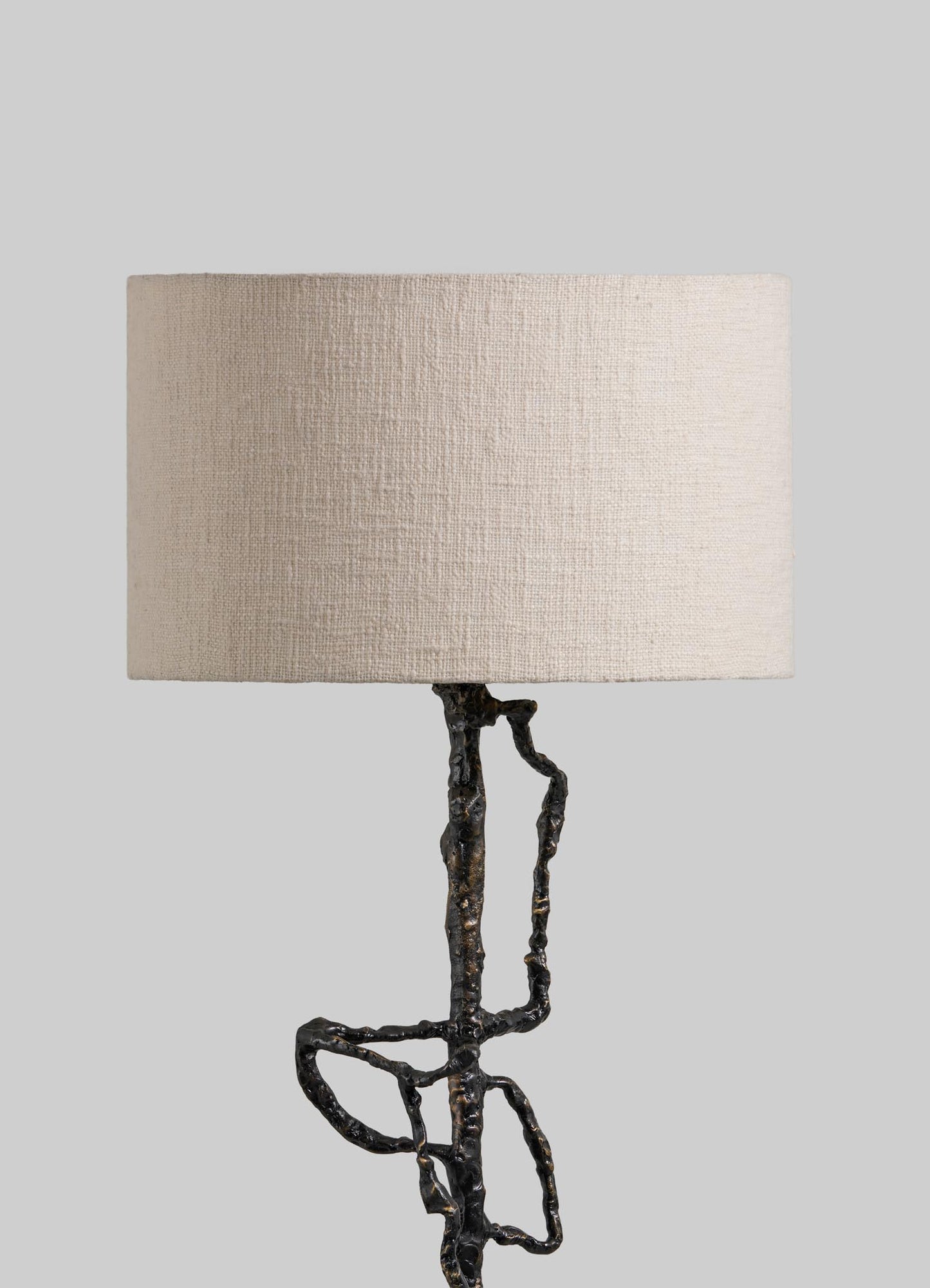 The VIYA Mohenjo Daro Floor Lamp features a beige textured cylindrical shade and an abstract black metal base inspired by Mohenjo-Daro artistry, displayed against a plain gray background.