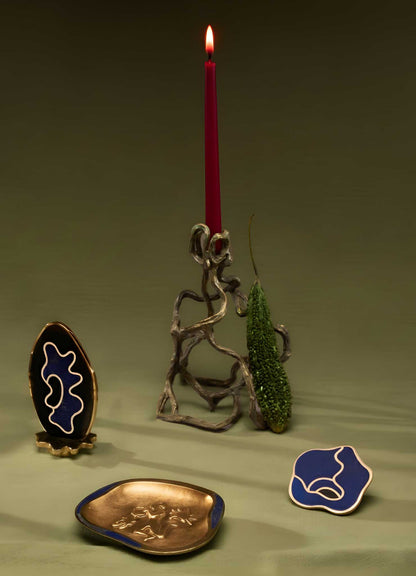 A VIYA Mohenjo Daro Candle Holder Medium glows with a red candle beside a green moss accent. In front, gold and blue decor pieces echo relics from Mohenjo-Daro, all arranged on a vivid green surface.