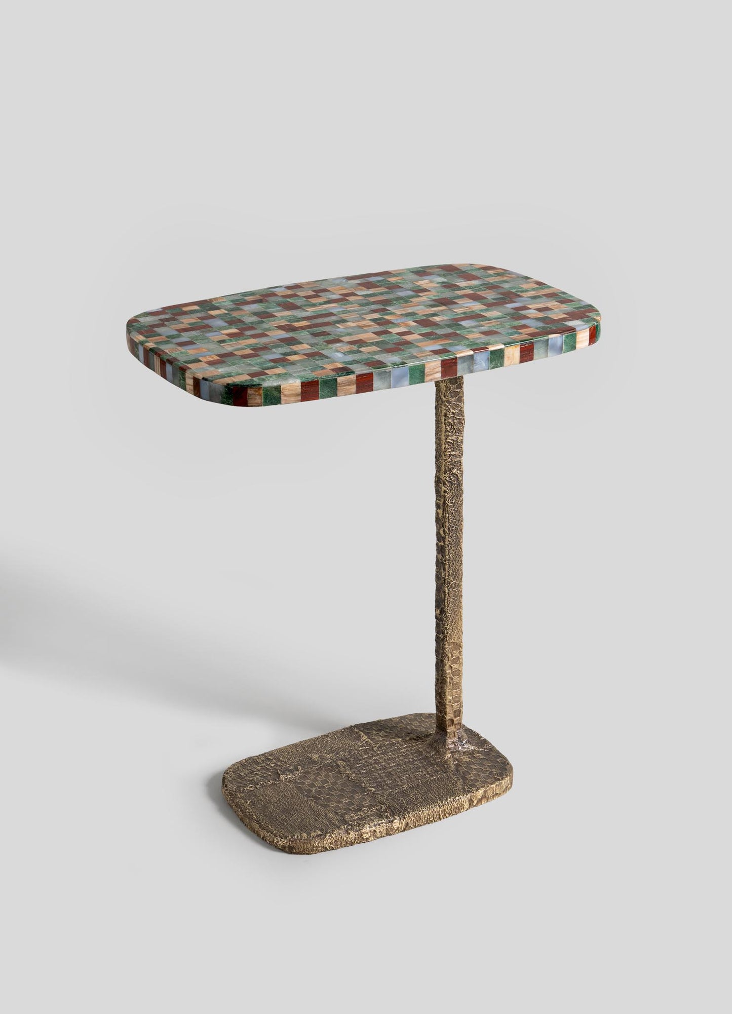 The VIYA Mosaic Side Table B features a colorful mosaic-patterned rectangular top made of semi-precious stones, with a single textured gold support and matching oval cast brass base, shown against a plain light gray background.