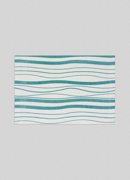 The Viya Madras Check Aqua Placemats (Set of 4) feature a white background with teal wavy horizontal lines in varying thicknesses, creating an abstract, flowing pattern inspired by Indian artistry. Shown centered on a light gray backdrop.