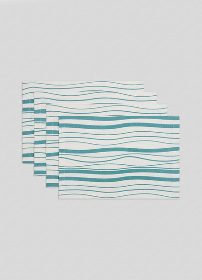 The Viya Madras Check Aqua Placemats (set of 4) feature a white base with wavy teal, light blue, and gray lines, arranged slightly overlapping on a plain light gray surface—a modern look inspired by the Indian subcontinent.