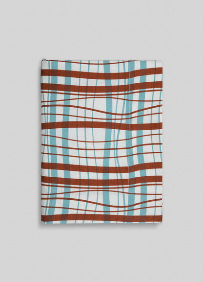 The Viya Madras Check Aqua & Rust Table Cloth showcases a folded fabric with an aqua and rust distorted plaid pattern, featuring curved and wavy lines, set against a light gray background.