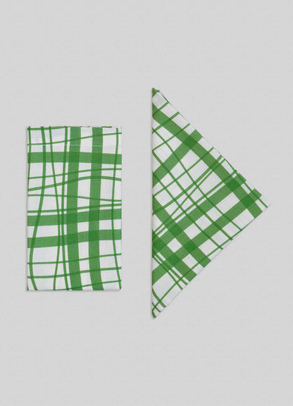 Two Viya Madras Check Green Napkins from the set of 4—one folded rectangular, one triangular—display Indian-inspired plaid patterns on a light grey background.
