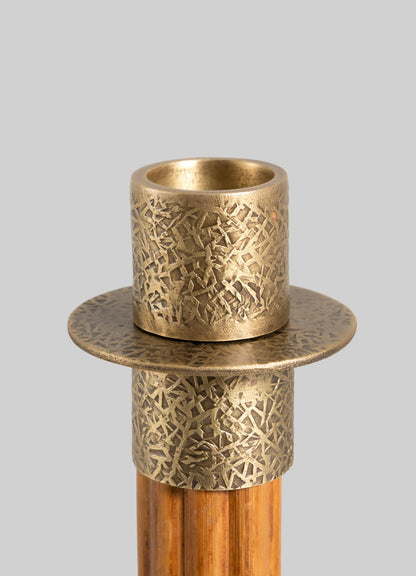 The VIYA Namsai Candle Holder Medium features a decorative brass design with intricate trimmings, a textured scratched finish, and a circular base on a cylindrical wooden stand, displayed against a plain gray background.