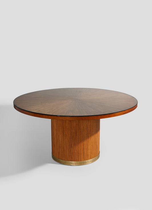 The VIYA Namsai Dining Table is a round wooden piece with a wide, flat top and cylindrical ribbed base, accented by refined brass trimmings at the bottom for an elegant touch.