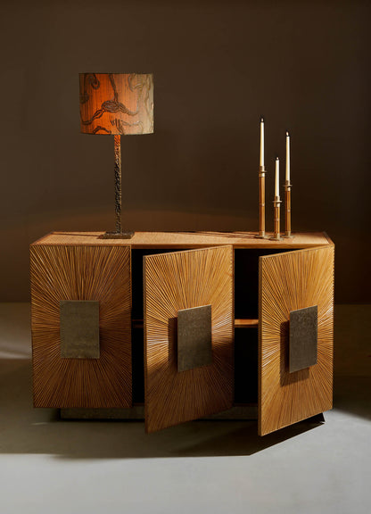 The VIYA Namsai Cabinet features a sunburst wooden design and brass handles, shown with doors open against a neutral background. It is topped with a textured-shade lamp and two tall candlesticks.