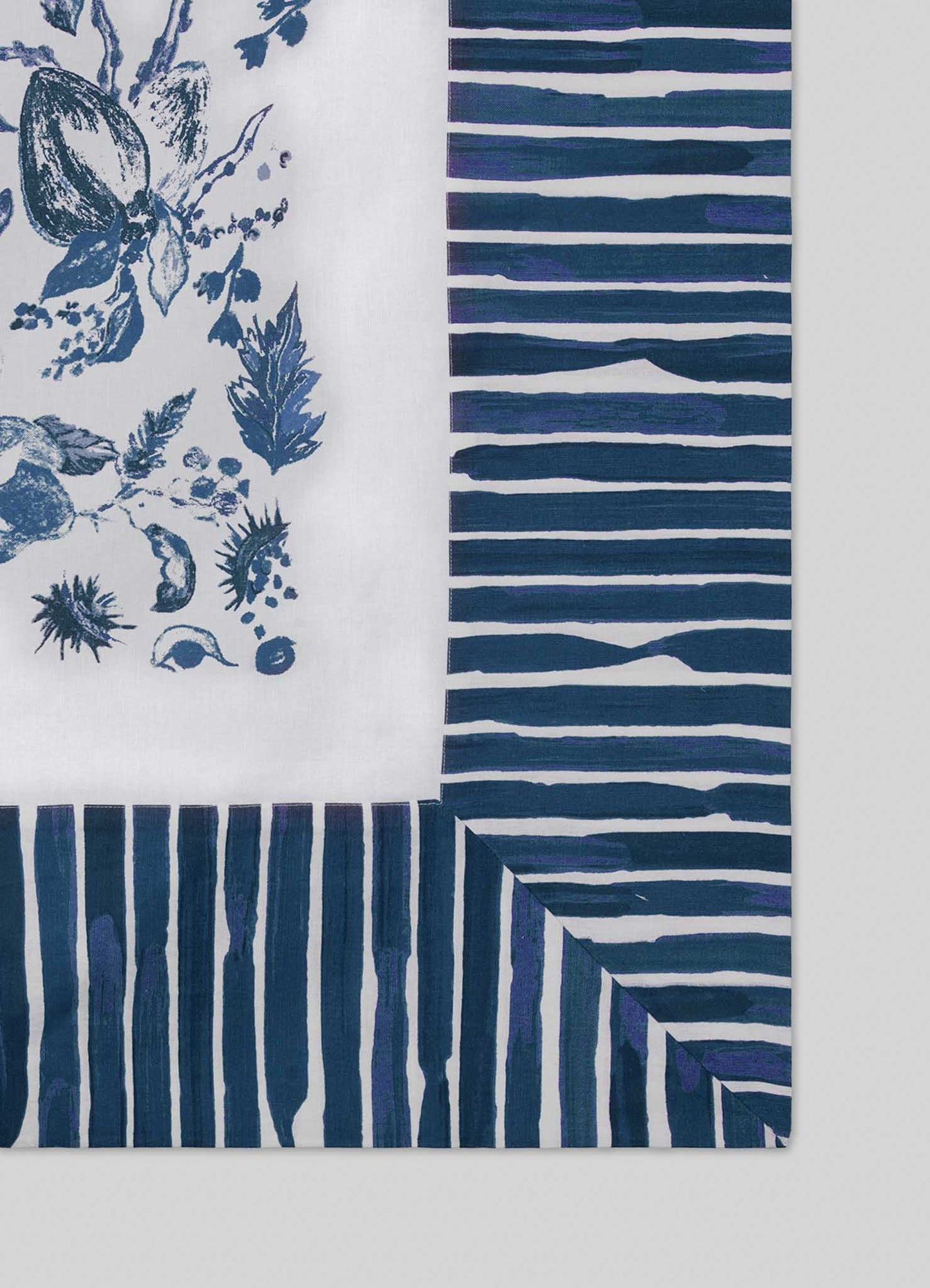 The Viya Orchard Blue Table Cloth Large features a white center with blue botanical designs inspired by Indian orchards, bordered by hand-painted blue and white stripes running along the edges.