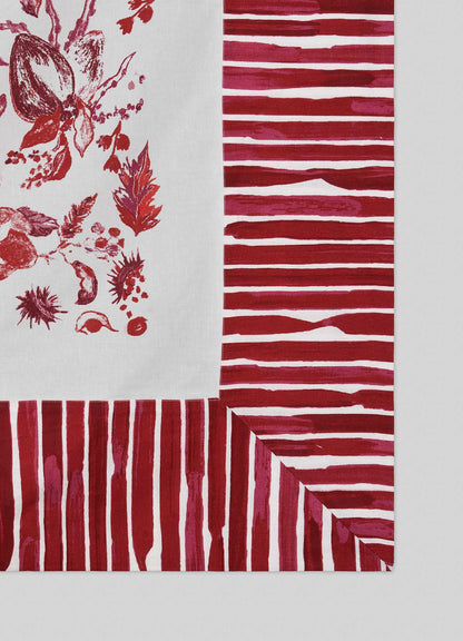 A close-up of Viya's Orchard Red Table Cloth Large shows a white fabric corner with red botanical prints inspired by Indian pomegranate orchards, accented by borders of alternating red stripes in hand-painted patterns.