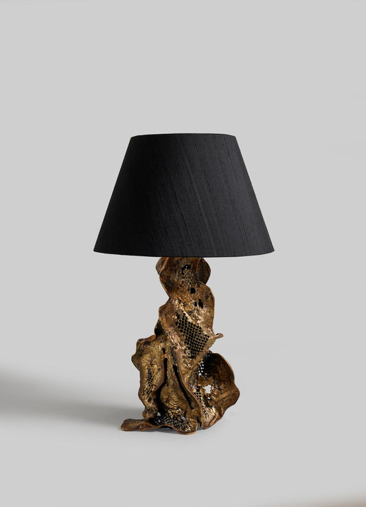 The VIYA Palash Table Lamp features a sculptural, twisted metallic base inspired by driftwood and a wide black shade, shown against a plain light grey background.