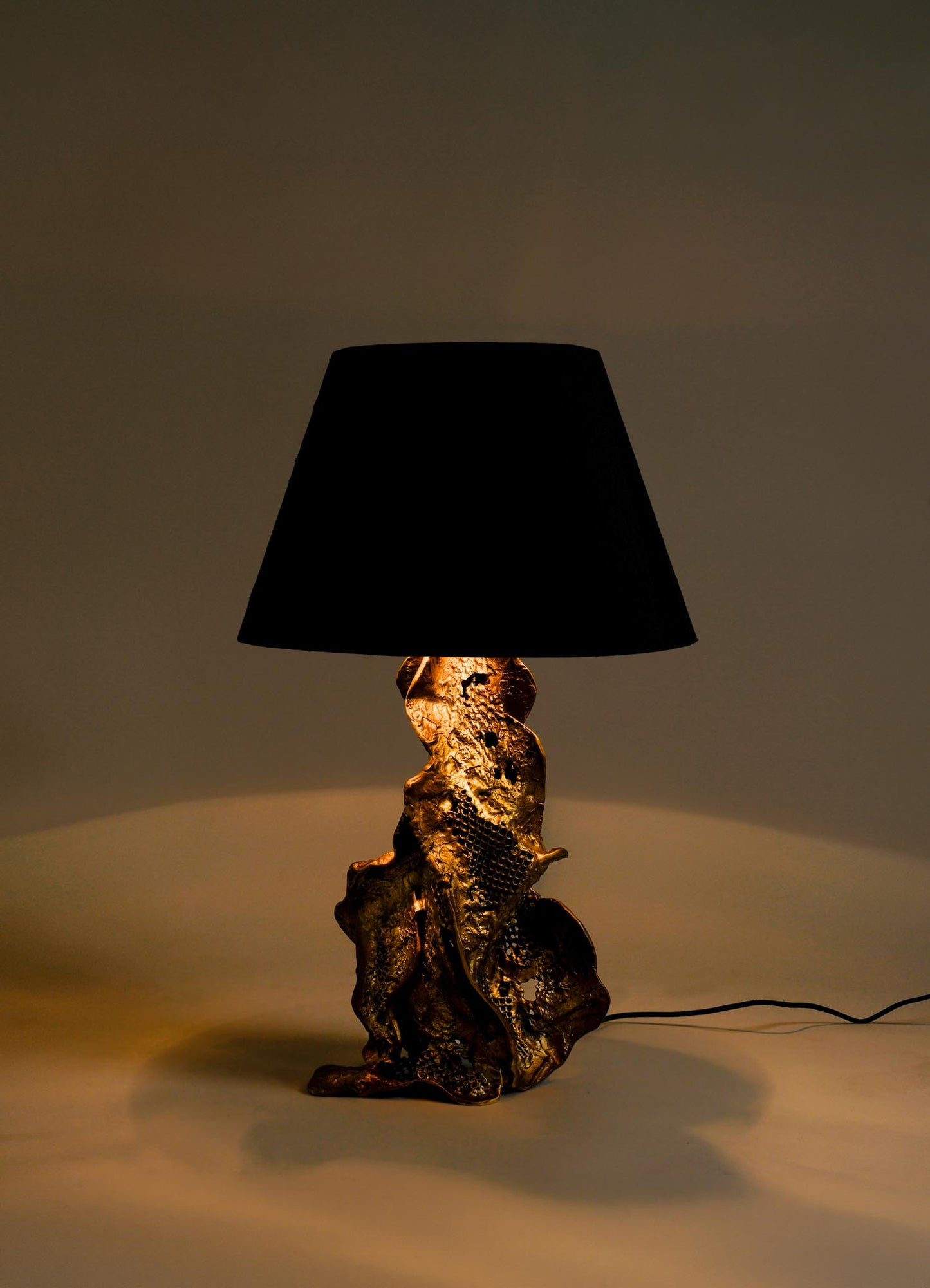 The VIYA Palash Table Lamp features a sculpted, textured golden base with abstract shapes and a large black lampshade, softly illuminating any space against a plain background.