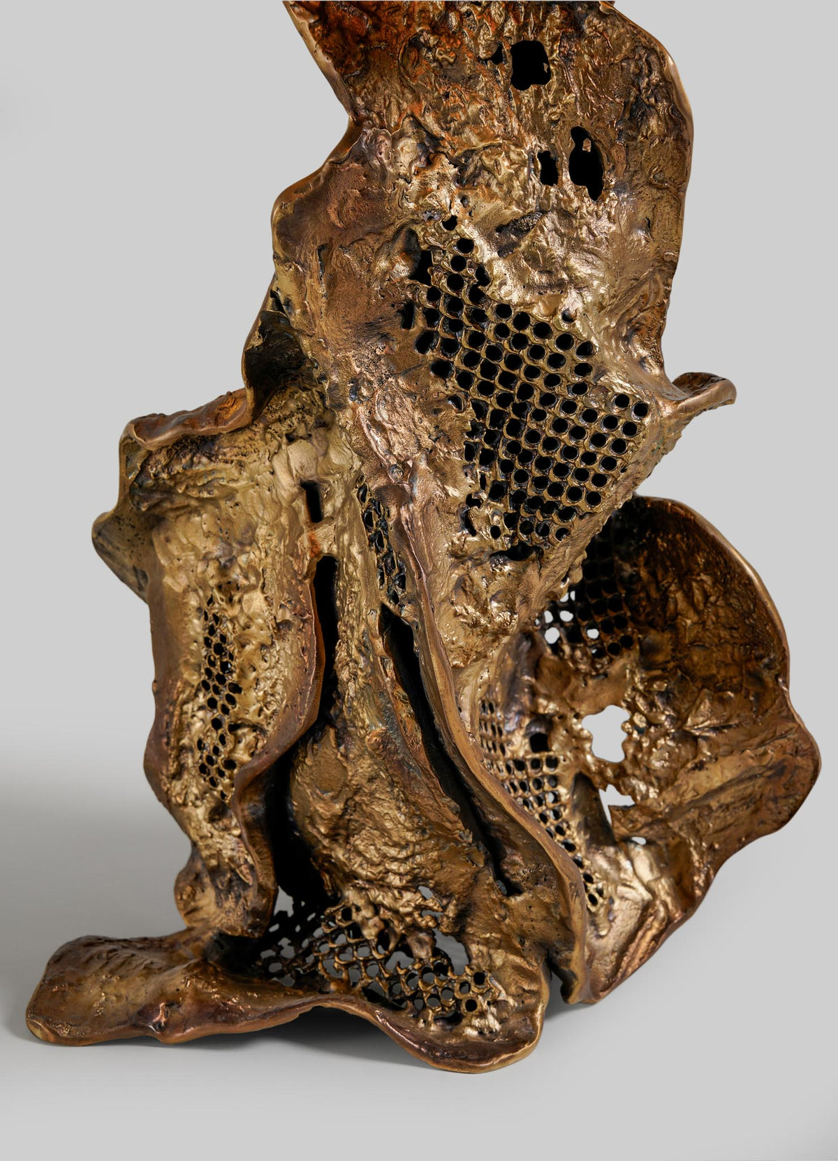 The Palash Table Lamp by VIYA features an abstract bronze design with organic, flowing shapes and honeycomb-patterned perforations. Its textured surface sits on a flat base, set against a plain gray background.