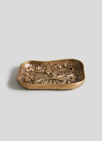 The VIYA Tiger & Hare Trinket Tray, a rectangular bronze piece with wavy edges and embossed animal and floral designs, makes a striking accent, styled on a plain light grey background.