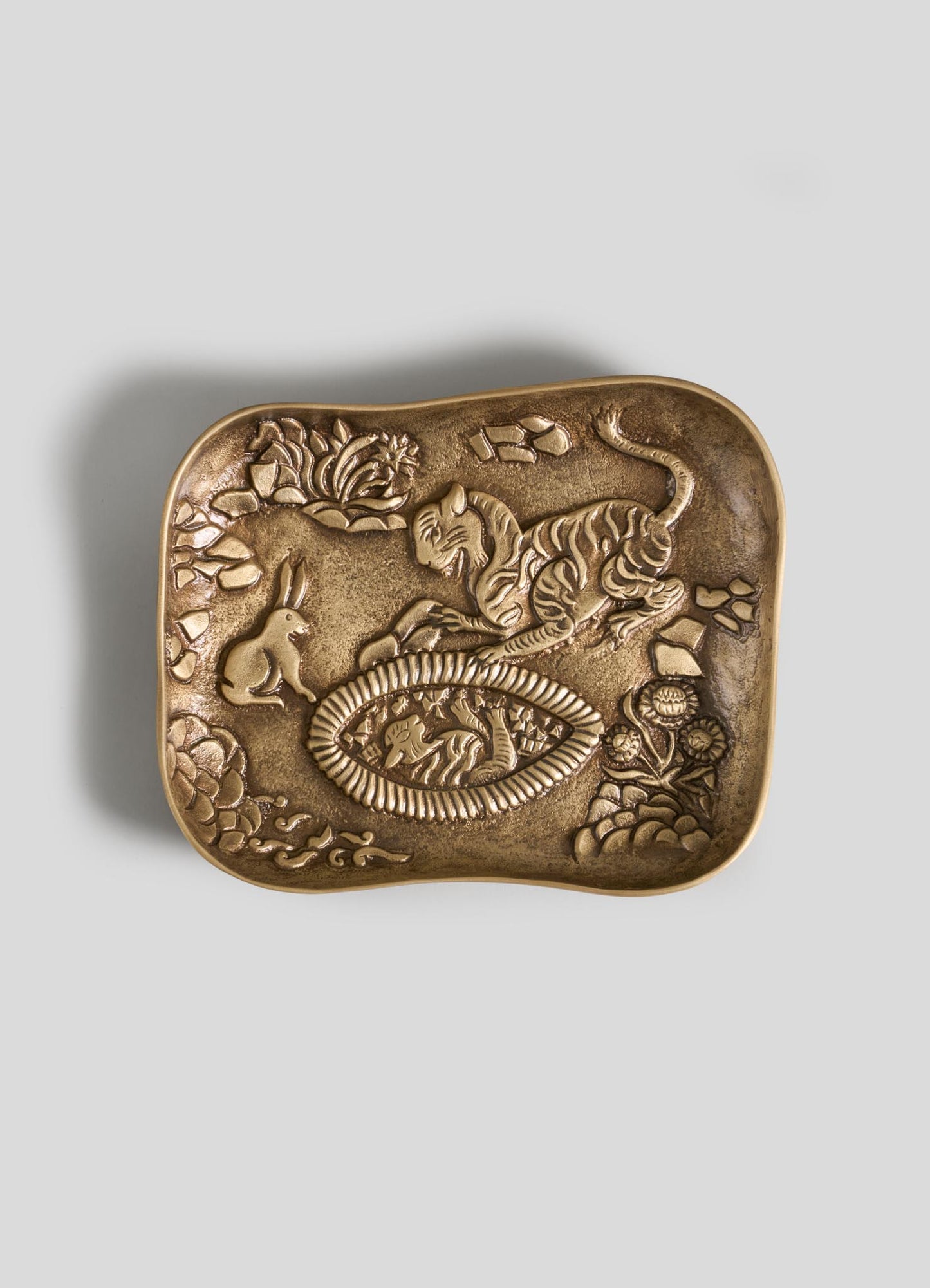 The VIYA Tiger & Hare Trinket Tray is a rectangular bronze tray with a textured surface, embossed jungle-inspired designs, and slightly curved edges. It features a tiger, rabbit, and flowers around a central oval pattern.
